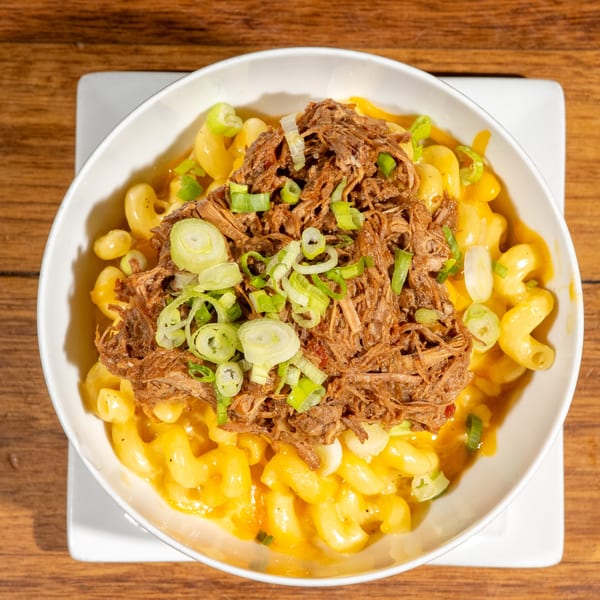 A white bowl of macaroni and cheese topped with shredded beef and green onions on a wooden table.