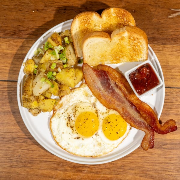 A breakfast plate with two fried eggs, crispy bacon, home fries with scallions, toast, and jam.